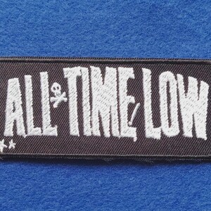 May include: A rectangular black embroidered patch featuring the white text ALL TIME LOW in a stylised font. The design includes a small white skull and crossbones icon between the words and two small white stars in the bottom left corner.