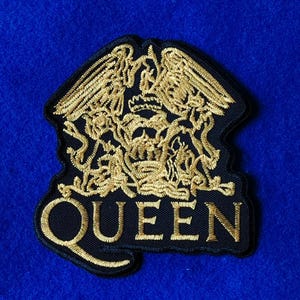 May include: A black embroidered patch featuring the gold Queen band crest logo. The design includes a central crown, two lions, a crab, and two fairies, all rendered in metallic gold thread. The word QUEEN is stitched in gold at the bottom of the patch.