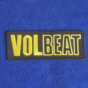 May include: A rectangular embroidered patch featuring a black background with the word VOLBEAT stitched in bold, yellow capital letters. The patch has a clean, stitched border and is designed for application onto fabric surfaces.