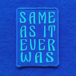 Peut inclure: Un écusson rectangulaire brodé avec un fond bleu et une bordure bleu clair. Le texte SAME AS IT EVER WAS est brodé en lettres capitales bleu clair au centre de l'écusson, avec une police de caractères stylisée et légèrement incurvée.