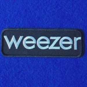 May include: A rectangular embroidered patch with a black background and the word weezer stitched in white lowercase letters. The patch features a thick black border and a textured fabric surface, displaying the band name in a clean font.