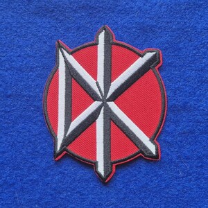 May include: A circular embroidered patch featuring a red background with a black and white stylised logo design. The emblem consists of a vertical bar intersected by diagonal lines, creating a geometric shape with sharp, pointed edges.