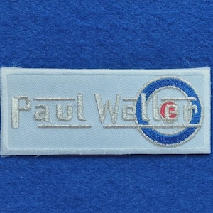 以下が含まれることがあります： 白い背景に銀色の糸で「Paul Weller」という文字が刺繍された長方形のワッペン。文字の右側には、青と白の同心円状のリングと中央に小さな赤いディテールが施された円形のロゴがあり、すべて糸で刺繍されています。