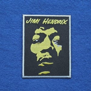 May include: A rectangular embroidered patch featuring a black background with a yellow stylised portrait of Jimi Hendrix. The name JIMI HENDRIX is embroidered in yellow text at the top. The patch has a white border and a black stitched edge.