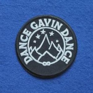 May include: A circular black embroidered patch featuring white stitching. The text DANCE GAVIN DANCE curves around the border. The centre displays a mountain range graphic with four small stars above the peaks and an infinity symbol below.