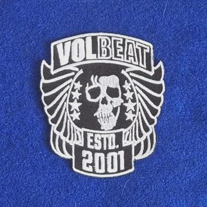 May include: A black and white embroidered patch featuring a central skull graphic flanked by wings and stars. The text VOLBEAT is arched at the top, with ESTD. 2001 printed at the bottom. The design is enclosed in a shield-like border.