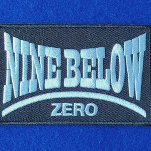 May include: A rectangular embroidered patch with a black background and light blue stitching. The text NINE BELOW is arched across the top, with the word ZERO centred below it, separated by a curved light blue line.