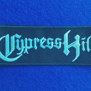 May include: A rectangular embroidered patch featuring the text Cypress Hill in a light blue gothic-style font. The lettering is stitched onto a black fabric background with a black merrowed border, creating a high-contrast design.