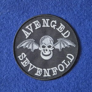 May include: A circular black embroidered patch featuring a white skull with bat wings in the centre. The text "AVENGED" is arched above the skull, and "SEVENFOLD" is arched below the skull in white capital letters with a black border.
