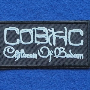 Könnte beinhalten: Ein rechteckiger, schwarz bestickter Aufnäher mit weißem Text. Die obere Zeile zeigt die Buchstaben COBHC in einer stilisierten Schriftart, während die untere Zeile die Worte Children Of Bodom in Schreibschrift mit einem schwarzen Rand enthält.