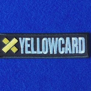 May include: A rectangular embroidered patch with a black background featuring a yellow cross-shaped icon on the left and the word YELLOWCARD in light blue capital letters on the right, all enclosed within a thin black border.