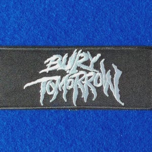 May include: A rectangular black embroidered patch featuring the band name Bury Tomorrow in a stylised, jagged white font. The embroidery has a textured finish with a distinct border, showcasing the band logo clearly against the black fabric background.