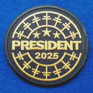 May include: A circular embroidered patch with a black background and gold thread detailing. The design features three stars above the word PRESIDENT, with the year 2025 below it, all enclosed within a circular border with radiating crossbar lines.