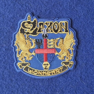 Könnte beinhalten: Ein gestickter Aufnäher mit dem Wort SAXON in goldener Schrift oben und LIONHEART unten. Das Design zeigt zwei goldene Greifen, die ein zentrales Schild mit einem roten Kreuz, einem Speer und einem schwarzen Dreizack auf blauem Grund flankieren.