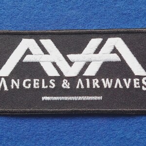 Peut inclure: Un écusson rectangulaire brodé noir présentant les lettres stylisées blanches AVA en haut et le texte ANGELS & AIRWAVES en dessous en lettres capitales blanches. Une petite ligne horizontale cousue blanche est centrée sous le texte.