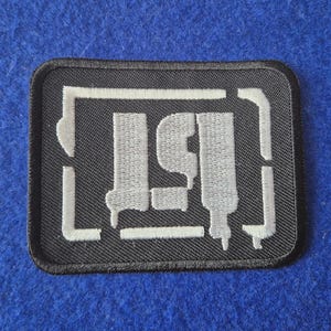 May include: A rectangular embroidered patch featuring a black background with a white, stylised graphic design. The design consists of a central abstract shape resembling a frame or symbol with dripping paint effects, all rendered in white thread.