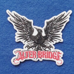 May include: An embroidered patch featuring a black bird with wings spread wide, positioned above the text "ALTER BRIDGE" in red, stylised lettering. The entire design is outlined in white thread, creating a distinct border against the background.
