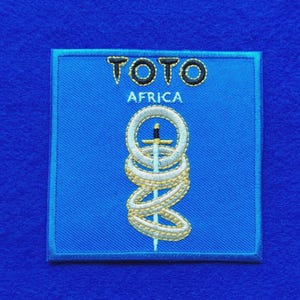 Può includere: Una toppa ricamata quadrata con sfondo blu brillante. Il testo TOTO e AFRICA è cucito con filo nero e bianco. Sotto il testo, una spada argentata è raffigurata verticalmente, intrecciata da tre anelli bianchi con accenti dorati.