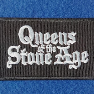 May include: A rectangular black embroidered patch featuring the white text Queens of the Stone Age in a stylised gothic font. The patch has a black stitched border and is designed for application to fabric surfaces like clothing or bags.