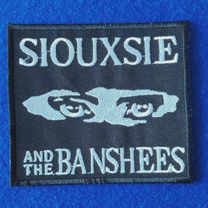 May include: A square black embroidered patch featuring the white text SIOUXSIE at the top and AND THE BANSHEES at the bottom. In the centre, there is a stylised white graphic illustration of two eyes with a jagged, abstract shape surrounding them.