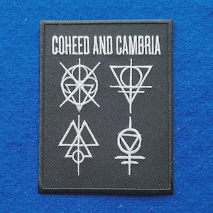 May include: A rectangular black embroidered patch featuring the white text COHEED AND CAMBRIA at the top. Below the text are four distinct white geometric symbols arranged in a two-by-two grid, each composed of intersecting lines, circles, and triangles.