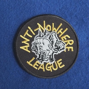 Op de afbeelding: Een ronde geborduurde patch met een zwarte achtergrond en een gele rand. De tekst ANTI-NOWHERE LEAGUE is in het geel gestikt rond een centrale wit-zwarte grafische illustratie van een gestileerde, grillige en getextureerde kop van een wezen.