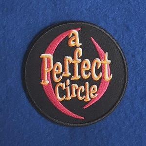 Puede incluir: Un parche bordado circular con fondo negro. El texto "a Perfect Circle" está cosido con hilo amarillo y naranja en el centro. Dos líneas bordadas curvas de color rosa enmarcan el texto, creando un elemento de diseño circular en el parche.