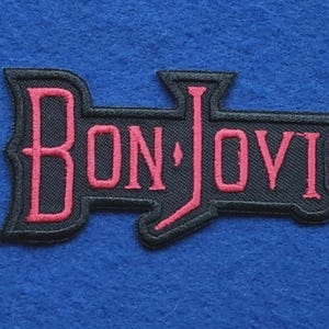 Może przedstawiać: Prostokątna haftowana naszywka z napisem BON JOVI wykonanym pogrubioną, różową, stylizowaną czcionką. Litery są obszyte czarną nicią na czarnym tle, z wyraźną obwódką dopasowaną do kształtu napisu.