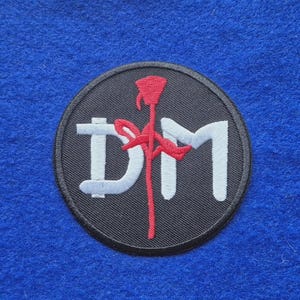 May include: A circular embroidered patch featuring a black background with a white letter D and a white letter M. A red rose with a long stem is positioned vertically between the letters, with the stem crossing over the D and the M.