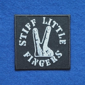 Peut inclure: Un écusson carré brodé noir présentant une main blanche faisant un signe de paix au centre. Le texte STIFF LITTLE FINGERS est brodé en lettres capitales blanches suivant un motif circulaire autour du graphique de la main sur le fond noir.