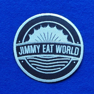 Puede incluir: Un parche bordado circular con fondo negro y costuras blancas. El diseño muestra un sol estilizado saliendo sobre las olas con el texto JIMMY EAT WORLD centrado en el medio en una fuente blanca, negrita y sin remates.