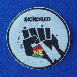 Può includere: Una toppa ricamata circolare con sfondo bianco e bordo nero. Presenta la parola SKINDRED in testo nero sopra un grafico nero di un pugno alzato. All'interno del pugno c'è il testo RAGGA THE POWER in rosso, giallo e verde.