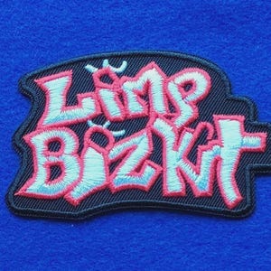 May include: An embroidered patch featuring the text Limp Bizkit in a stylised font. The lettering is light blue with a thick pink outline, set against a solid black background. The patch has a black stitched border and a jagged, irregular shape.