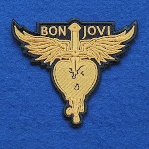 Puede incluir: Un parche bordado que presenta un corazón de color dorado atravesado por una espada con alas que se extienden desde la empuñadura. El texto BON JOVI está cosido en oro sobre las alas. El diseño está delineado en hilo negro sobre fondo azul.