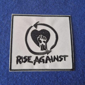 May include: A square embroidered patch with a white background and a black border. The design features a black heart with a raised fist inside, surrounded by a circular arrow. The text RISE AGAINST is embroidered in black below the icon.