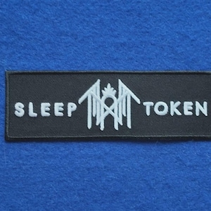 Op de afbeelding: Een rechthoekige zwarte geborduurde patch met de witte tekst SLEEP TOKEN en een centraal wit gestileerd grafisch symbool bestaande uit hoekige lijnen en een kroonachtige vorm tussen de twee woorden op een zwarte stoffen achtergrond.