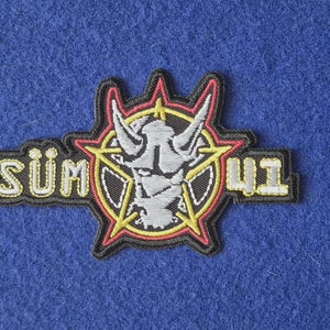 May include: An embroidered patch featuring the text SUM 41 in yellow and white lettering. The design includes a central grey horned figure enclosed within a yellow star and a red circular border, all outlined in black thread on a dark background.