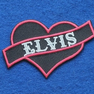 May include: An embroidered patch featuring a black heart shape outlined in pink thread. A diagonal black banner crosses the heart, displaying the name ELVIS in white capital letters. The entire design is bordered with a pink stitched edge.