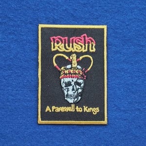 May include: A rectangular embroidered patch with a black background and a gold border. It features the word RUSH in pink and yellow, a crown graphic above a skull, and the text A Farewell to Kings in yellow thread at the bottom.