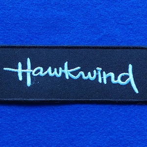 Puede incluir: Un parche rectangular negro bordado con la palabra Hawkwind cosida en hilo azul claro. El texto está escrito en una fuente cursiva estilizada en el centro del parche, que tiene un borde negro y una textura de tela visible.