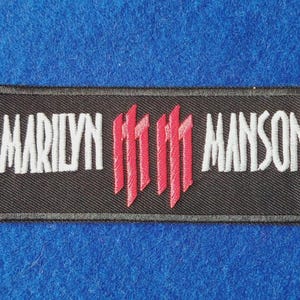 Op de afbeelding: Een rechthoekige geborduurde patch met een zwarte achtergrond. De tekst MARILYN MANSON is in witte hoofdletters aan de linker- en rechterkant geborduurd. In het midden staan twee verticale rode symbolen, elk bestaande uit drie parallelle lijnen.