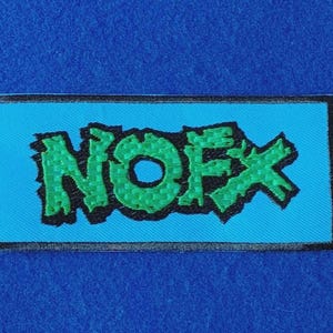 May include: A rectangular embroidered patch featuring the text NOFX in a jagged, bright green font with a thick black outline. The lettering is set against a solid, vibrant light blue background, all enclosed within a thin black border.