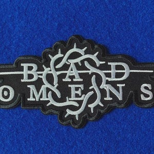 May include: A black embroidered patch featuring the text BAD OMENS in white capital letters. The design includes a central white circular emblem of intertwined thorny vines that intersect with a horizontal line running through the text.