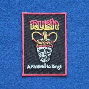 May include: A rectangular embroidered patch with a black background and a red border. It features the word RUSH in yellow and red, a crown resting on a skull, and the text A Farewell to Kings in white thread at the bottom.