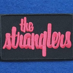 May include: A rectangular black embroidered patch featuring the text "the stranglers" in bright pink thread. The lettering is in a stylised, rounded font, and the entire patch is bordered by a thin black stitched edge, creating a clear contrast.
