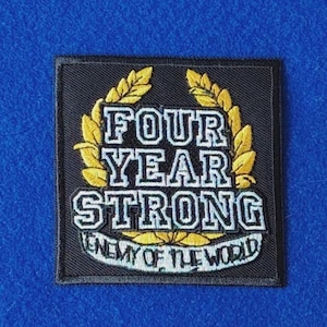May include: A square black embroidered patch featuring a yellow laurel wreath surrounding the white text FOUR YEAR STRONG. Below this, a white banner contains the text ENEMY OF THE WORLD in black lettering, all set against a blue background.