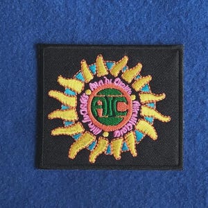 Könnte beinhalten: Ein quadratischer, schwarzer Stickaufnäher mit einem zentralen grünen Kreis, der die roten Buchstaben AIC enthält. Der Kreis ist von einem Ring aus rosa Text und einem Sonnenmotiv mit gelben Strahlen und blauen Akzenten umgeben.