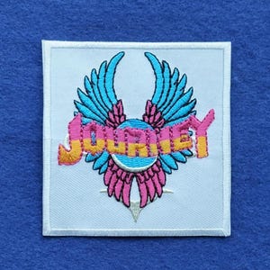 May include: A square white embroidered patch featuring the word Journey in pink and yellow letters. The text is layered over a circular blue emblem with stylised blue and pink wings extending upwards and downwards from the centre of the design.
