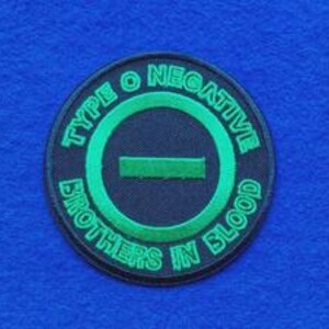 May include: A circular embroidered patch featuring a black background with a vibrant green border and centre horizontal bar. The text "TYPE O NEGATIVE" is arched across the top and "BROTHERS IN BLOOD" is arched across the bottom in green.