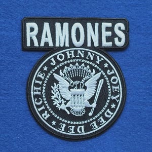May include: A black embroidered patch set featuring a rectangular top piece with the white text RAMONES and a circular bottom piece displaying a white eagle emblem surrounded by the names Johnny, Joey, Dee Dee, and Richie in white lettering.